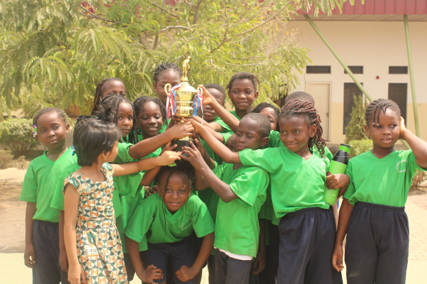 Eine Gruppe von Kindern hält einen Pokal in einem Schulhof mit Bäumen, Pflanzen und einem Gebäude im Hintergrund.