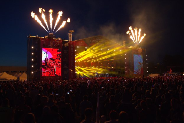 Große Menschenmenge auf einem Musikfestival mit beleuchteter Bühne, Performern, Bannern, Zelten links und Feuerwerk am dunklen Himmel.