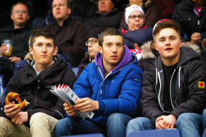 Eine Gruppe von Menschen sitzt im Stadion, einige tragen Jacken und Mützen, einer hält ein Objekt und ein anderer ein Getränk, und ein Mann mit einem menschlichen Gesicht in der Mitte.