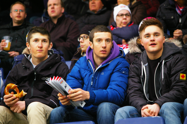 Eine Gruppe von Menschen sitzt im Stadion, einige tragen Jacken und Mützen, einer hält ein Objekt und ein anderer ein Getränk, und ein Mann mit einem menschlichen Gesicht in der Mitte.