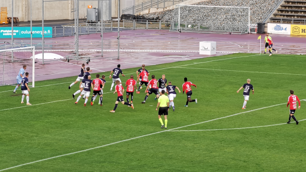 Eine Gruppe von Menschen, die Fußball auf einem Feld mit Zuschauern in einem Stadion spielen, umgeben von Pfosten, Tafeln mit Text, Geländern, Stufen und anderen Gegenständen.