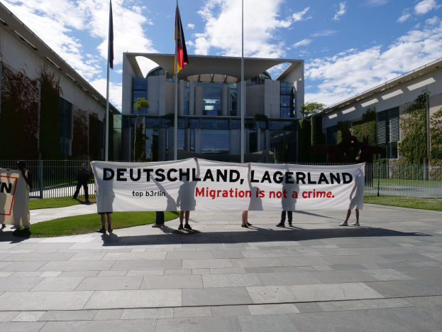 Eine Gruppe von Menschen mit einem Transparent mit der Aufschrift 'Deutschland, Lagerland Migration ist kein Verbrechen' vor einem Gebäude mit Fenstern, umgeben von Gras, einem Metallzaun, Pflanzen, Bäumen und Fahnenstangen unter einem bewölkten Himmel.