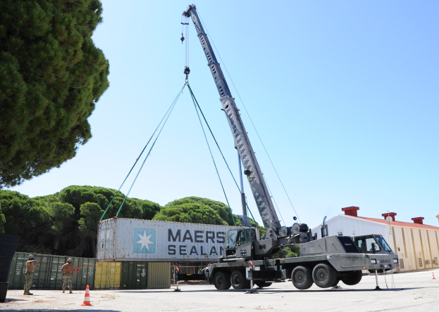 Ein Lastwagen mit einem Kran, der einen Container auf seine Ladefläche hebt, umgeben von Absperrgittern, Menschen, Containern, Bäumen und Gebäuden unter einem klaren blauen Himmel.