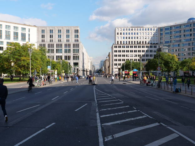 Eine belebte Straße in Berlin, Deutschland, mit Füttern und Radfahrern auf der Straße, hohen Gebäuden, Bäumen, Laternen und Schildern unter einem bewölktem Himmel.