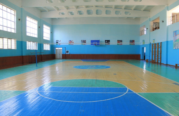 Eine große Turnhalle mit einem blauen und weißen Boden, die einen Basketballplatz, ein Netz, Stangen, eine Tür, Fenster, Wandplakate, ein Geländer und ein Dach zeigt.