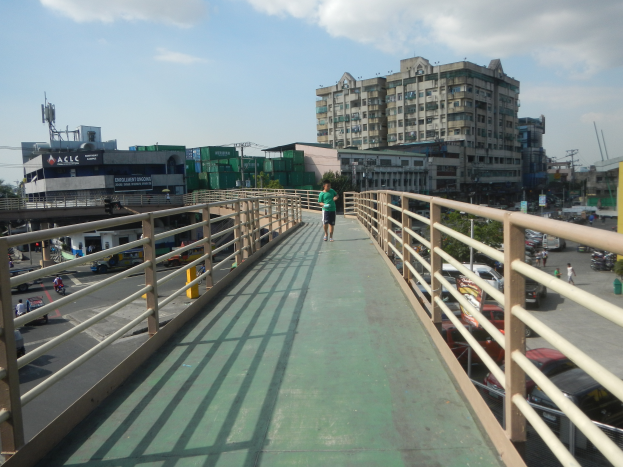 Ein Mann geht über eine Fussgängerbrücke über eine belebte Straße, mit Fahrzeugen darunter und Gebäuden, Bäumen und Polen im Hintergrund unter einem bewölktem Himmel.