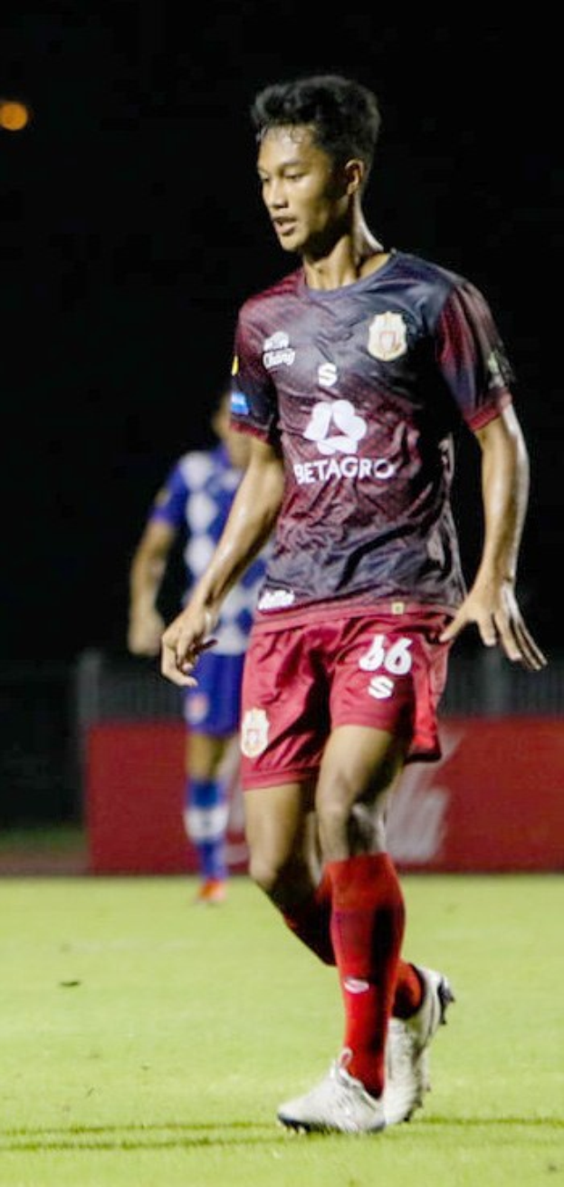 Ein Profifußballer in einem rotbraunen Trikot und roten Shorts, der einen Ball auf einem Feld kickt, mit einer anderen Person im Hintergrund und einer Tafel mit Text.