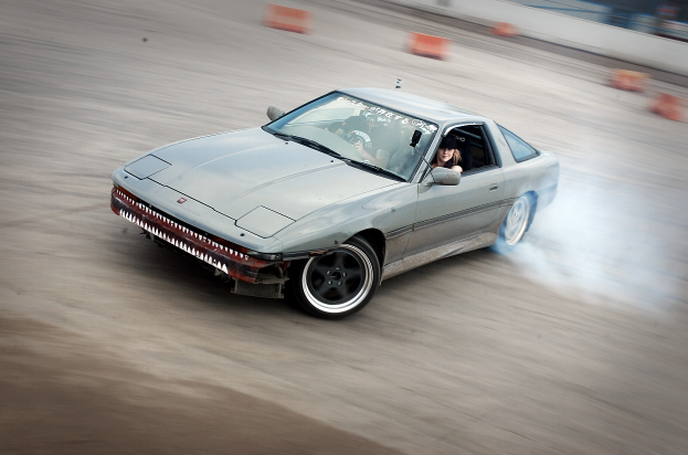 Eine Person in einem Helm fährt ein Auto auf einer Strecke, gibt Rauch ab, umgeben von orangefarbenen Gegenständen, mit einem unscharfen Hintergrund, der hohe Geschwindigkeit andeutet.