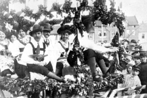 Schwarzes und weißes Bild eines Paradewagens, der Menschen in traditioneller bayrischer Tracht trägt, einige spielen Musikinstrumente, der Wagen ist mit Blättern und Dekorationen geschmückt, vor einem Hintergrund von Gebäuden unter einem klaren Himmel.