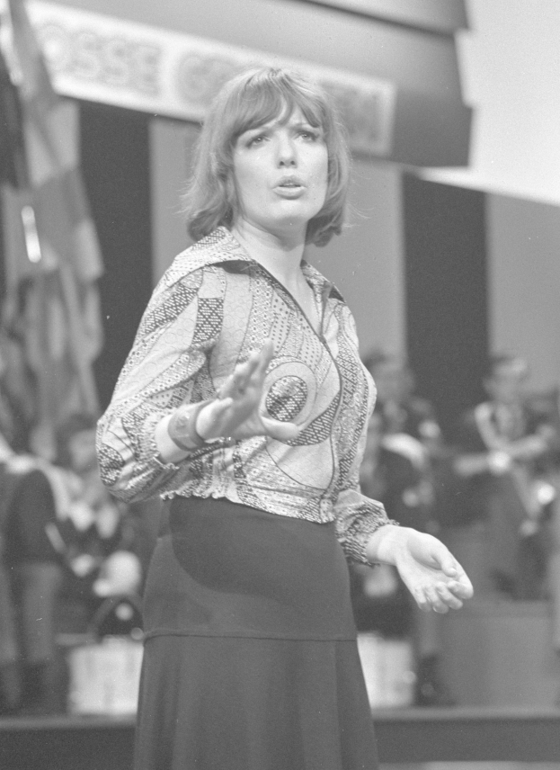 Eine Frau in einem Schwarz-Weiß-Foto steht auf einer Bühne, trägt ein weißes Hemd und einen schwarzen Rock, hat ihr Haar zurückgebunden und einen ernsten Gesichtsausdruck, umgeben von einer Gruppe von Menschen und einer Wand im Hintergrund.