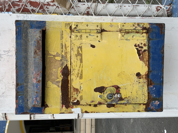 Ein gelb-blauer Metalltresor steht auf einer weißen Wand, im Hintergrund ist ein Metallzaun zu sehen.