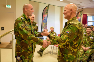 Zwei Männer in Militäruniformen geben sich die Hand vor einer sitzenden Gruppe, mit einem Tisch, Geländer, Bannern, einer Tür, einem Schild, einem Fenster mit Vorhang und Deckenleuchten im Hintergrund.