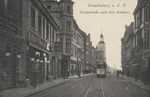 Ein altes Schwarz-Weiß-Foto einer Stadtstraße in Brandenburg, Deutschland, das eine Straßenbahn auf Schienen, Menschen auf dem Gehweg, Gebäude mit Fenstern, Schilder und Straßenlaternen unter einem bewölkten Himmel zeigt.