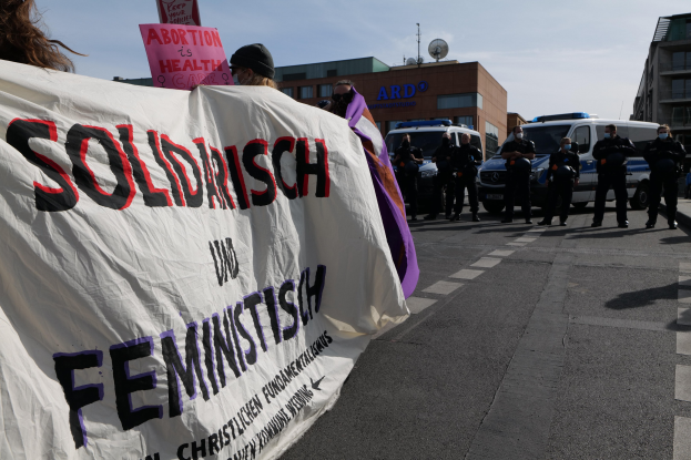 Eine Gruppe von Menschen marschiert auf einer Straße, hält eine Fahne mit der Aufschrift 'Solidarität und Feminismus', mit parkenden Fahrzeugen am Straßenrand und Gebäuden im Hintergrund unter einem klaren blauen Himmel.