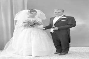 Ein Schwarz-Weiß-Foto eines lachenden Mannes und einer Frau, die nebeneinander auf ihrer Hochzeit stehen, mit der Frau in einem weißen Kleid und einem Blumenstrauß und einer Vorhangwand im Hintergrund.