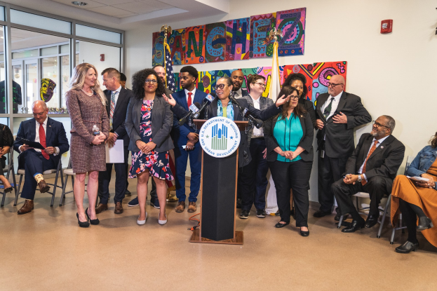 Eine Gruppe von Menschen steht vor einem Podium mit einem Mikrofon und einer Tafel mit der Aufschrift "Sancho City Councilman Joins City Council", während andere sitzen oder stehen; Flaggen und ein Feuerlöscher sind an der Rückwand zu sehen.