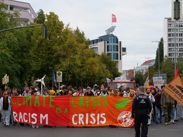 Eine Gruppe von Menschen marschiert eine von Bäumen gesäumte Straße entlang und hält ein Transparent mit der Aufschrift "Klima-Krise ist eine Krise", mit parkenden Fahrzeugen, Gebäuden und einem klaren blauen Himmel im Hintergrund.