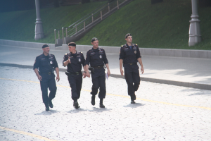 Drei Polizisten in Uniform und Mützen gehen eine Straße entlang mit Polen, Geländern, Treppen und Gras im Hintergrund.