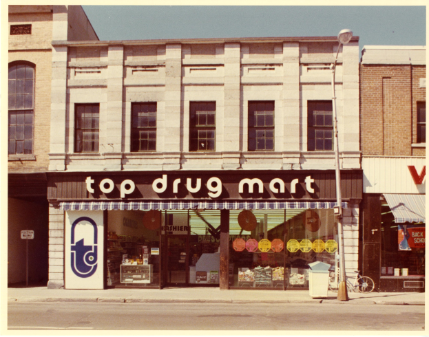 Top-Drogenmart-Filiale an einer Straßenecke mit einem Gebäude, Fenstern, einem Schild, einer Straßenlaterne, einem geparkten Fahrrad, einem Müllcontainer und einem bewölkten Himmel.