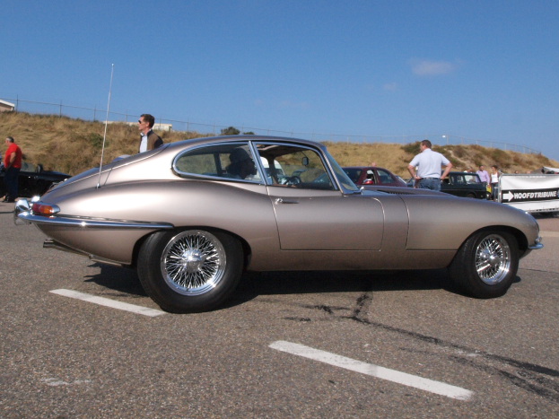 Ein silberfarbenes Auto auf einem Parkplatz mit Menschen in der Nähe, ein Banner im Hintergrund und ein bewölkter Himmel.