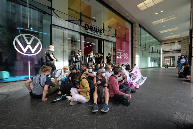 Menschen sitzen vor einer Volkswagen-Niederlassung auf dem Boden und halten ein Transparent während einer Demonstration.