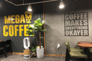 Außenansicht eines Kaffee-Shops namens "Mega Coffee" mit Tischen, Stühlen, einem Bodenreinigungsgerät, Deckenleuchten und einem Topfpflanze in Singapur.