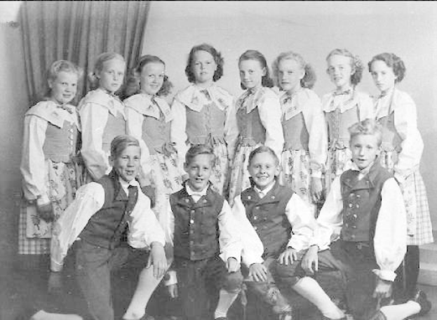 Schwarzes und weißes Foto einer Gruppe von Kindern, die vor einer Wand mit einem Vorhang posieren, mit Text unten, der "1930er-Kinderschule" lautet.
