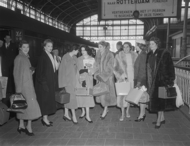 Schwarzes und weißes Bild von Frauen, die an einem Bahnhof stehen, mit einem Zug auf der linken Seite und Geländern und einer Tafel auf der rechten Seite, jede hält eine Tasche.