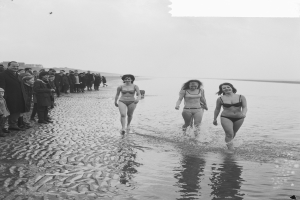 Eine Gruppe von Frauen in Bikinis, die an einem Strand entlanggehen, mit einigen Menschen auf der linken Seite, dargestellt in Schwarz-Weiß.