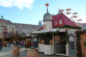 Ein lebendiger Weihnachtsmarkt in Nürnberg, Deutschland, mit Menschen um geschmückte Stände, Gebäuden, einem Riesenrad und einem bewölkten Himmel sowie einer Tafel auf der rechten Seite.
