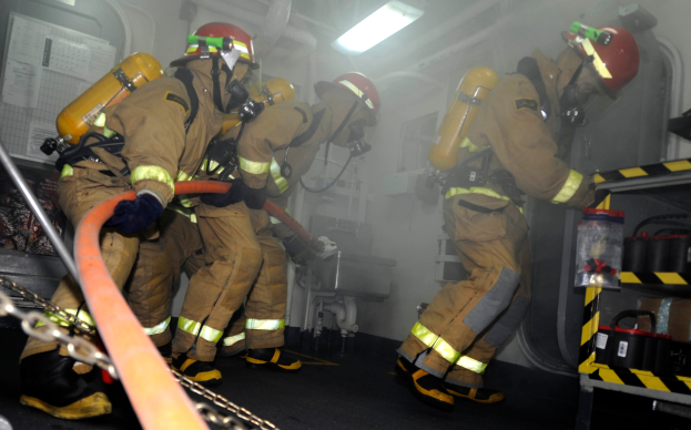 Eine Gruppe von Feuerwehrmännern in Schutzausröstung, darunter Helme und Sauerstoffflaschen, arbeitet an einem Feuerwehrauto. Rechts im Bild befindet sich ein Gestell mit verschiedenen Gegenständen und im Hintergrund Rohre, Lichter und andere Gegenstände.