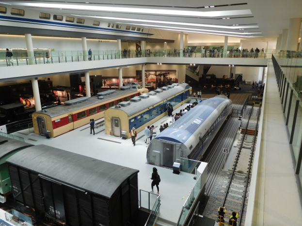Eine geräumige Museumsausstellung, die verschiedene Bahnen in unterschiedlichen Größen und Farben zeigt, mit Besuchern, die vom Boden, Geländern und Pfeilern unter Deckenleuchten beobachten.