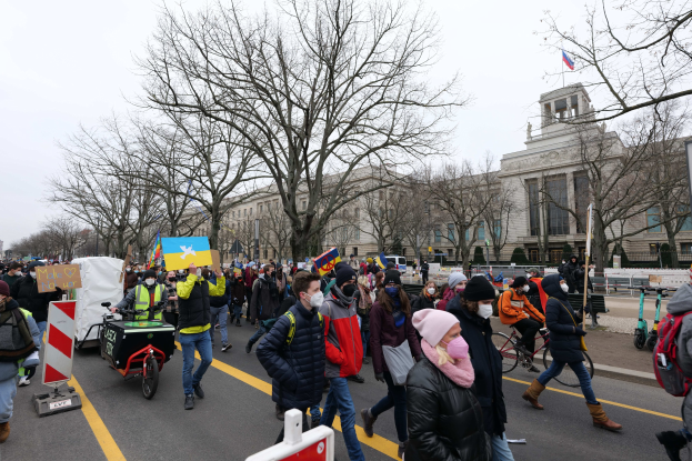 Eine große Gruppe von Menschen marschiert auf einer Straße in Washington, D.C. und hält Schilder und Banner hoch, während einige Fahrräder fahren, unter einem klaren blauen Himmel vor einem Gebäude.