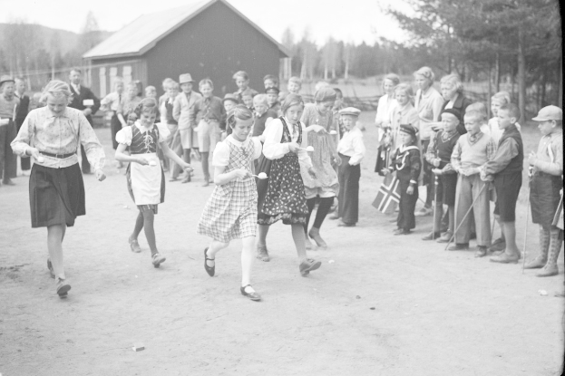 Schwarzes Foto von Kindern, die Fußball spielen, mit einer Menge von Menschen, die Fahnen halten, Bäumen und einem Haus im Hintergrund.