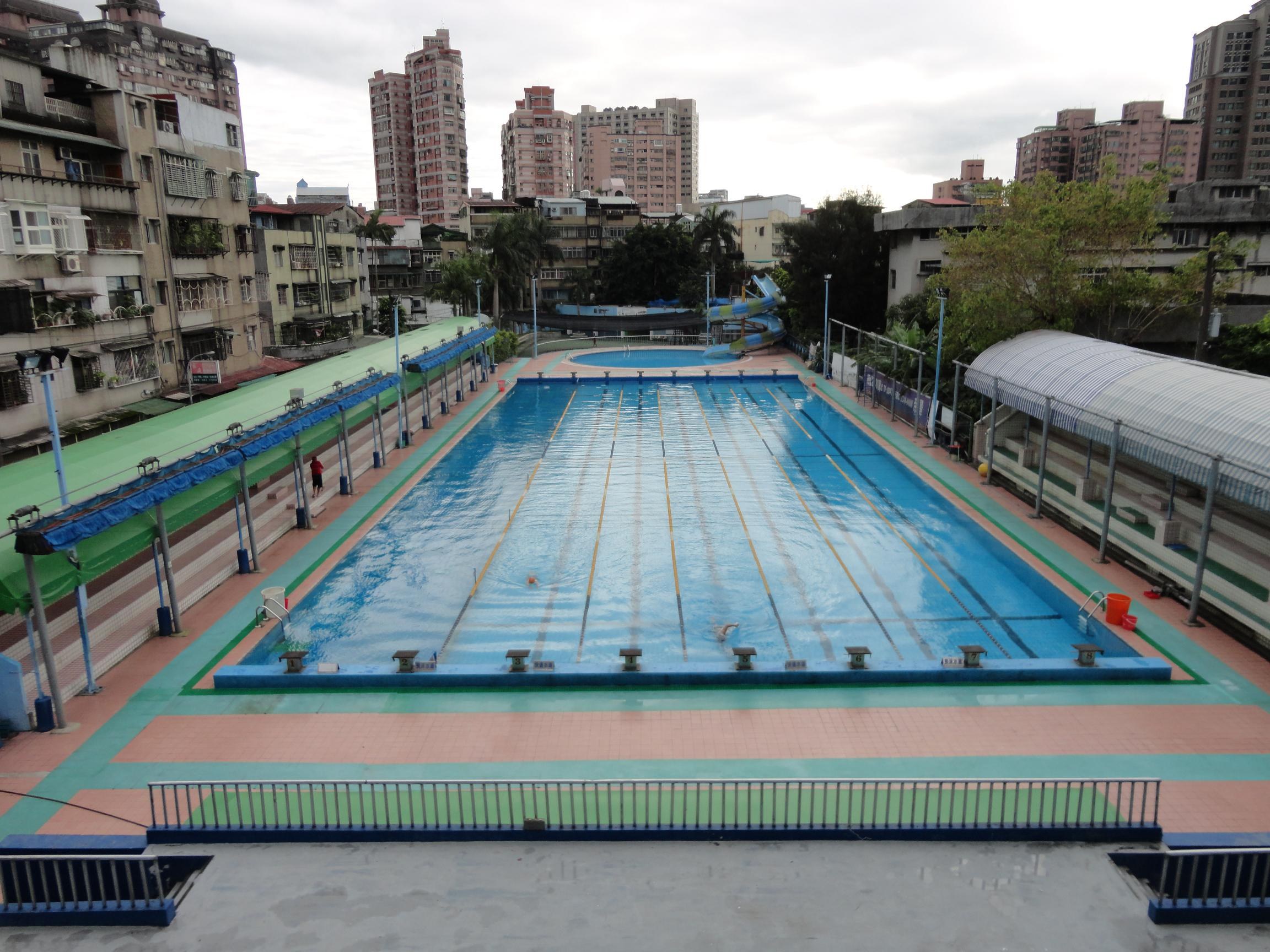 Großer städtischer Schwimmbadkomplex umgeben von Zäunen, Pfählen, Laternen, Bäumen und Gebäuden, mit einem Schuppen auf der rechten Seite und Himmel im Hintergrund.