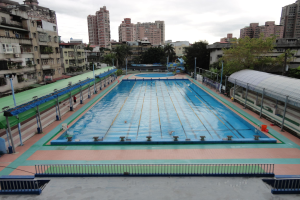 Großer städtischer Schwimmbadkomplex umgeben von Zäunen, Pfählen, Laternen, Bäumen und Gebäuden, mit einem Schuppen auf der rechten Seite und Himmel im Hintergrund.