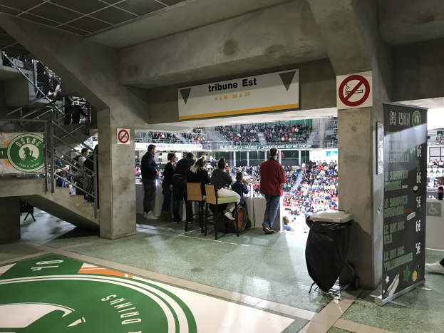 Gruppe von Menschen vor einer sitzenden Menge in einem Stadion mit Schildern, die "Boston Celtics vs New York Jets" lesen, einem Müllbehälter, einer Tafel mit Text, einer Treppe mit Geländern und einer Decke mit Deckenleuchten.