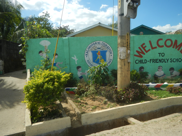 Eine grüne Wand mit einem Schild, das "Willkommen in der kinderfreundlichen Schule" trägt, umgeben von Häusern, Bäumen, Pflanzen und einem Mast mit einer Lampe, unter einem bewölkten Himmel.