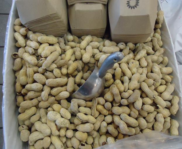 Eine Schachtel Erdnüsse auf einem Tisch mit einer Plastikabdeckung und einem Löffel daneben, umgeben von zusätzlichen Schachteln Erdnüsse.