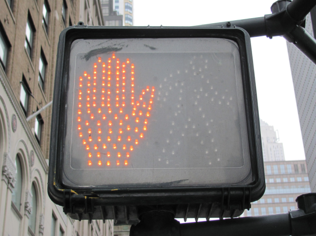Rote Hand auf dem Verkehrssignal in einer städtischen Umgebung mit hohen Gebäuden auf beiden Seiten und einem klaren Himmel darüber.