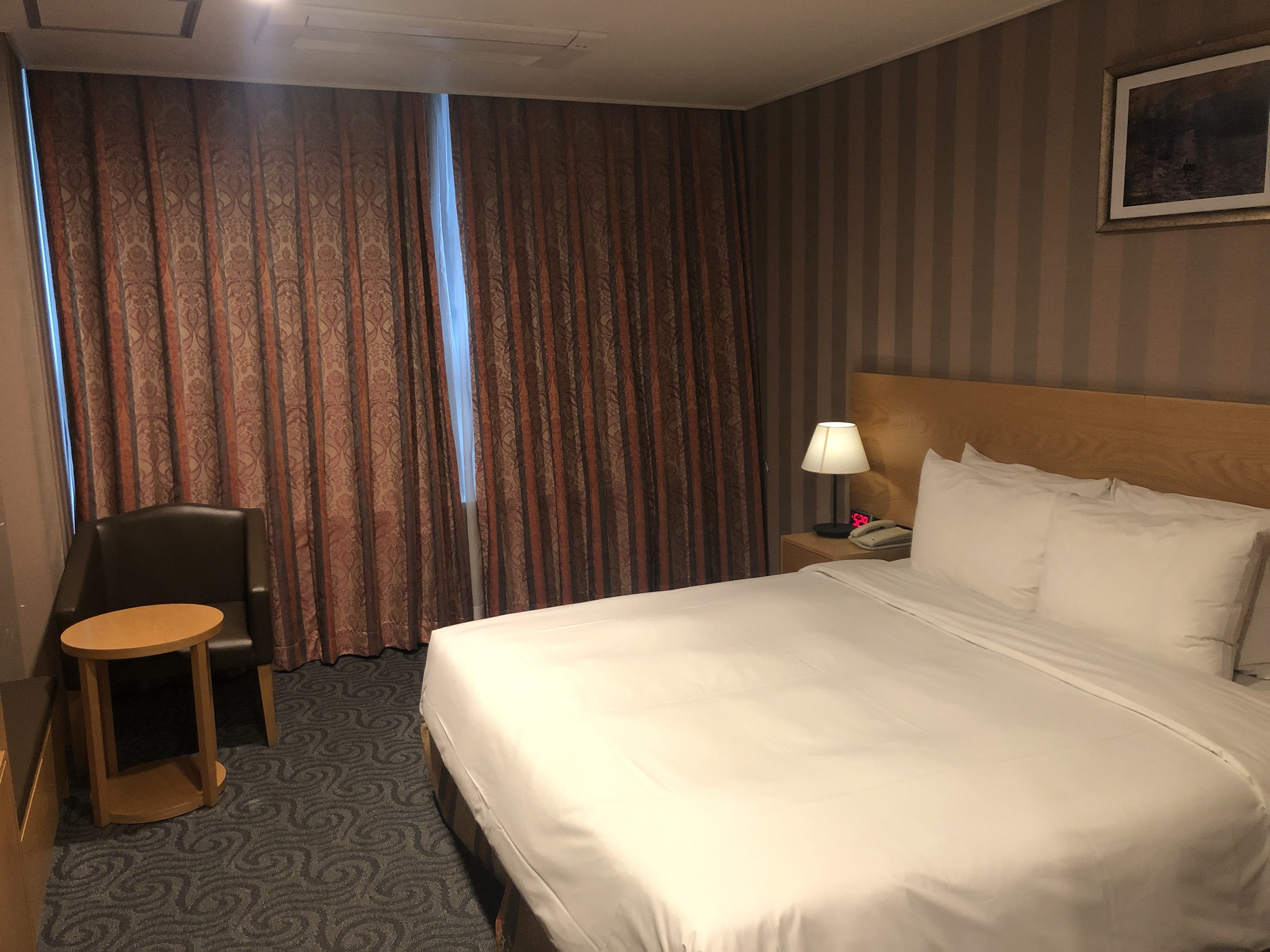 Hotelzimmer mit großem Bett, Stuhl, Tisch, Telefon, Lampe, Foto auf dem Wand, Fenster mit Vorhängen und Deckenbeleuchtung.