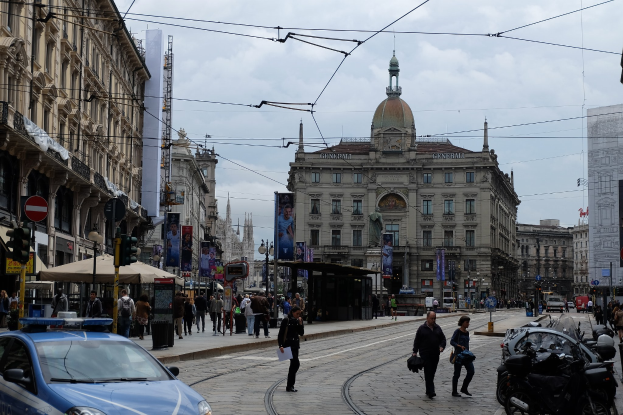 Eine belebte Straße mit einem parkenden Polizeiwagen, Passanten mit Taschen, fahrenden Fahrzeugen, Gebäuden mit Fenstern und Bannern, Laternenmästen, Verkehrsampeln und einem bewölktem Himmel.