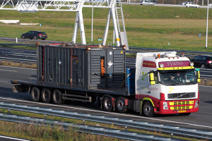 Ein Lkw mit einem großen Container fährt auf einer Autobahn, mit anderen Fahrzeugen, Pfählen, Bäumen, Gebäuden und einem klaren blauen Himmel im Hintergrund.
