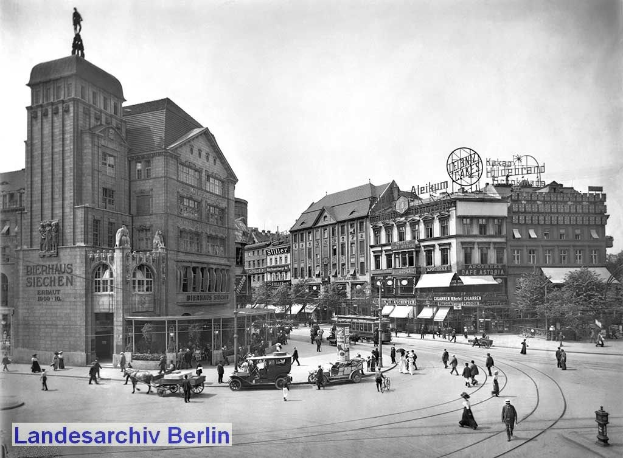 Schwarz-weiß-Foto einer belebten Berliner Stadtstraße mit Fußgängern, Fahrzeugen, einer Dachfigur, Beschilderung, Strommasten, Bäumen und einem bewölkten Himmel.