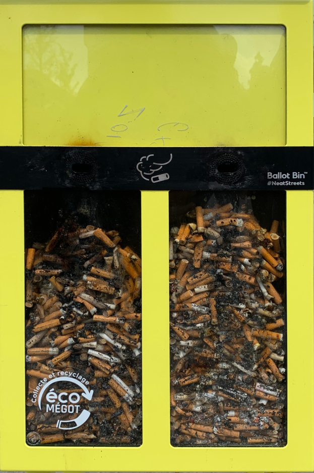 Ein leuchtend gelber Recyclingmüllcontainer voller Zigarettenstummeln auf einem Gehweg, beschriftet für die Wiederverwertung.