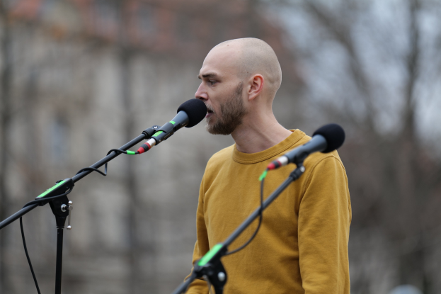 Ein Mann mit Glatze in einem gelben T-Shirt singt in ein Mikrofon mit einem unscharfen Hintergrund.