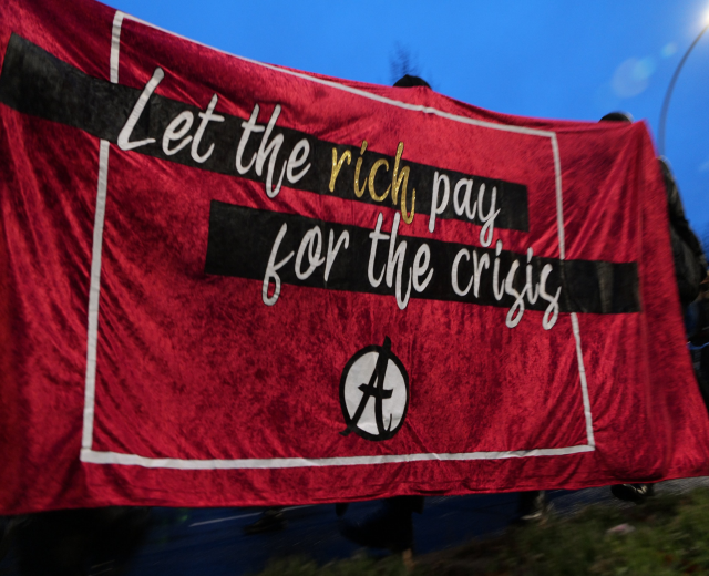 Gruppe von Menschen, die eine Straße entlanggehen und ein rotes Banner mit der Aufschrift "Lasst die Reichen für die Krise bezahlen" tragen, vor Bäumen und einem Laternenmast unter einem klaren blauen Himmel.