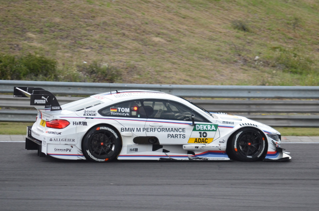 BMW M4 DTM-Rennwagen auf einer Rennstrecke mit Text auf dem Fahrzeug, umgeben von grünem Gras und einer Absperrung im Hintergrund.