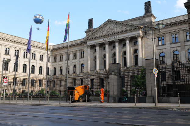 Großes modernes Regierungsgebäude mit vielen Fenstern, umgeben von Straßenmöbeln und Fahrzeugen, mit Menschen auf dem Bürgersteig und Fahnen davor.
