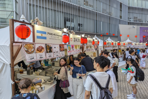 Eine Gruppe von Menschen auf einem Food-Festival in Japan, die um einen Tisch mit Essen versammelt sind, einige tragen Taschen und halten Gläser, während andere die Gegenstände auf dem Tisch untersuchen; Tafeln mit Text und Bildern, Lichter und Laternen hängen von der Decke, mit einem Gebäude im Hintergrund.